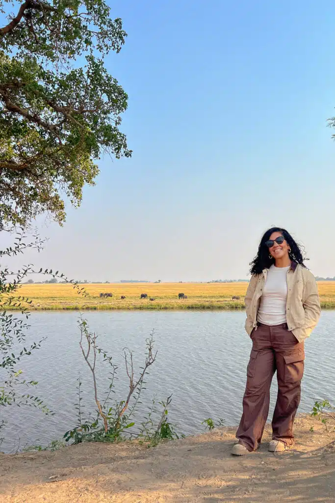 A woman in sunglasses stands by a riverbank, smiling with her hands in her pockets. Behind her, African Overland Tours reveal grassy plains with grazing animals beneath a clear blue sky, while tree branches frame the top left corner.