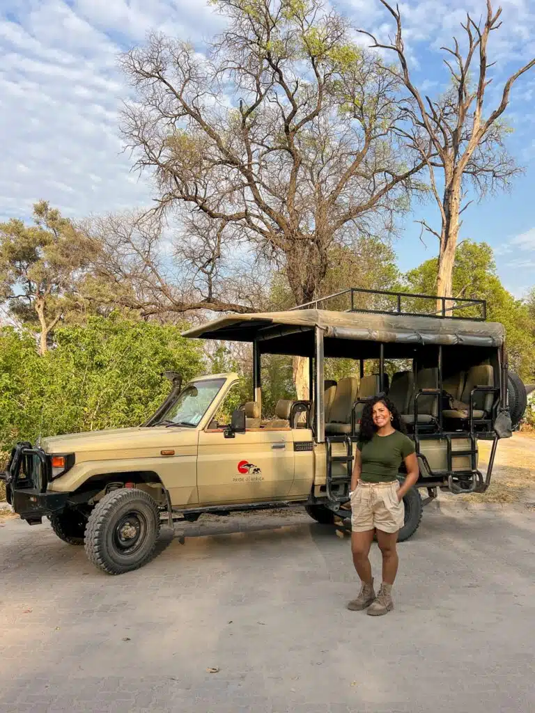 A woman in safari attire stands smiling in front of an open-sided safari vehicle, ready for one of the African Overland Tours, with greenery and tall trees in the background under a partly cloudy sky.