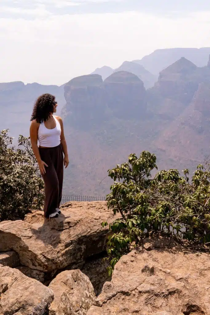 A woman with curly hair, wearing a white tank top and brown pants, stands on a rocky ledge overlooking a scenic canyon—a breathtaking view reminiscent of landscapes seen on African Overland Tours.
