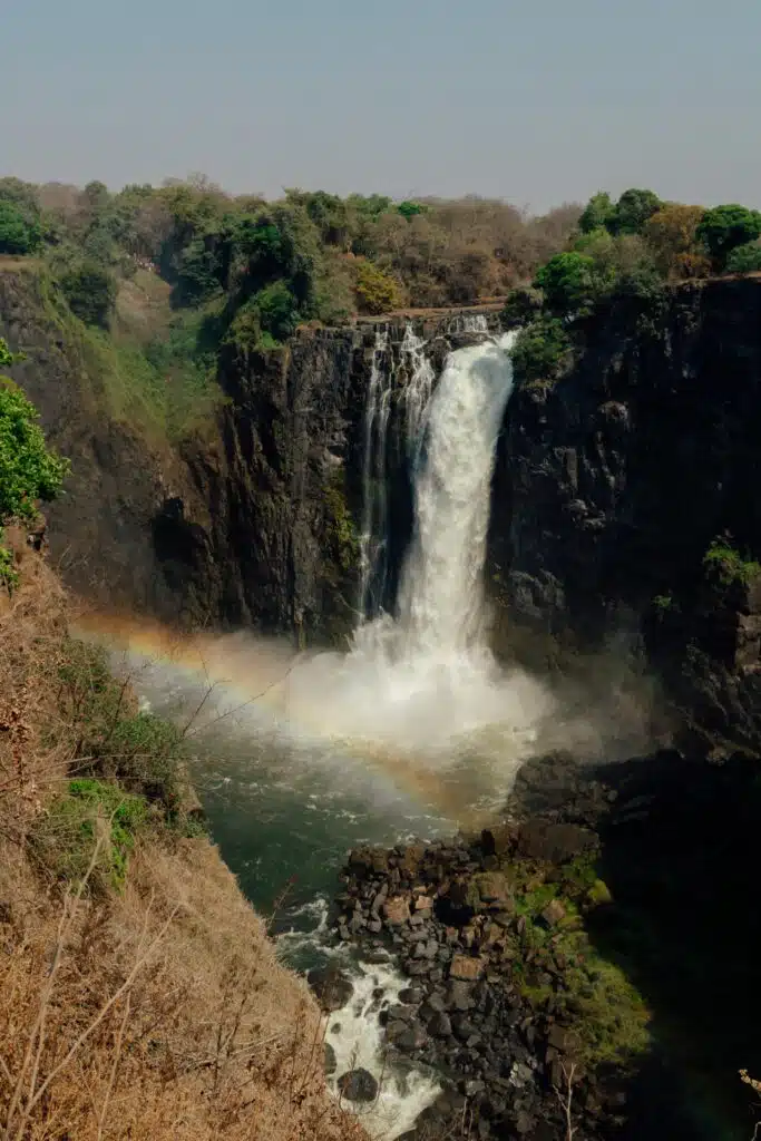 A tall waterfall cascades down a rocky cliff surrounded by lush green trees, with a rainbow forming in the mist at the base—a breathtaking sight often experienced on African Overland Tours.