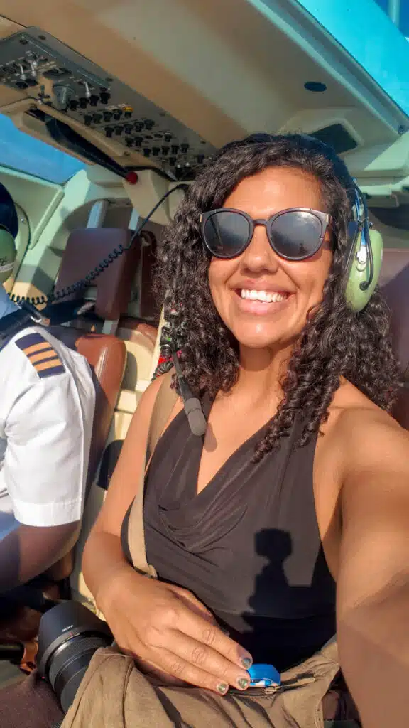A woman wearing sunglasses and a headset smiles for a selfie inside a helicopter during one of her African Overland Tours. Next to her, a pilot in uniform is partially visible as sunlight fills the cockpit.