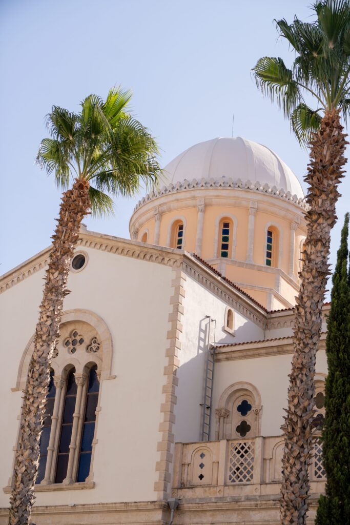 A white historic building with arched windows and a large domed roof is framed by two tall palm trees under a clear blue sky, capturing the charm of Limassol in winter.