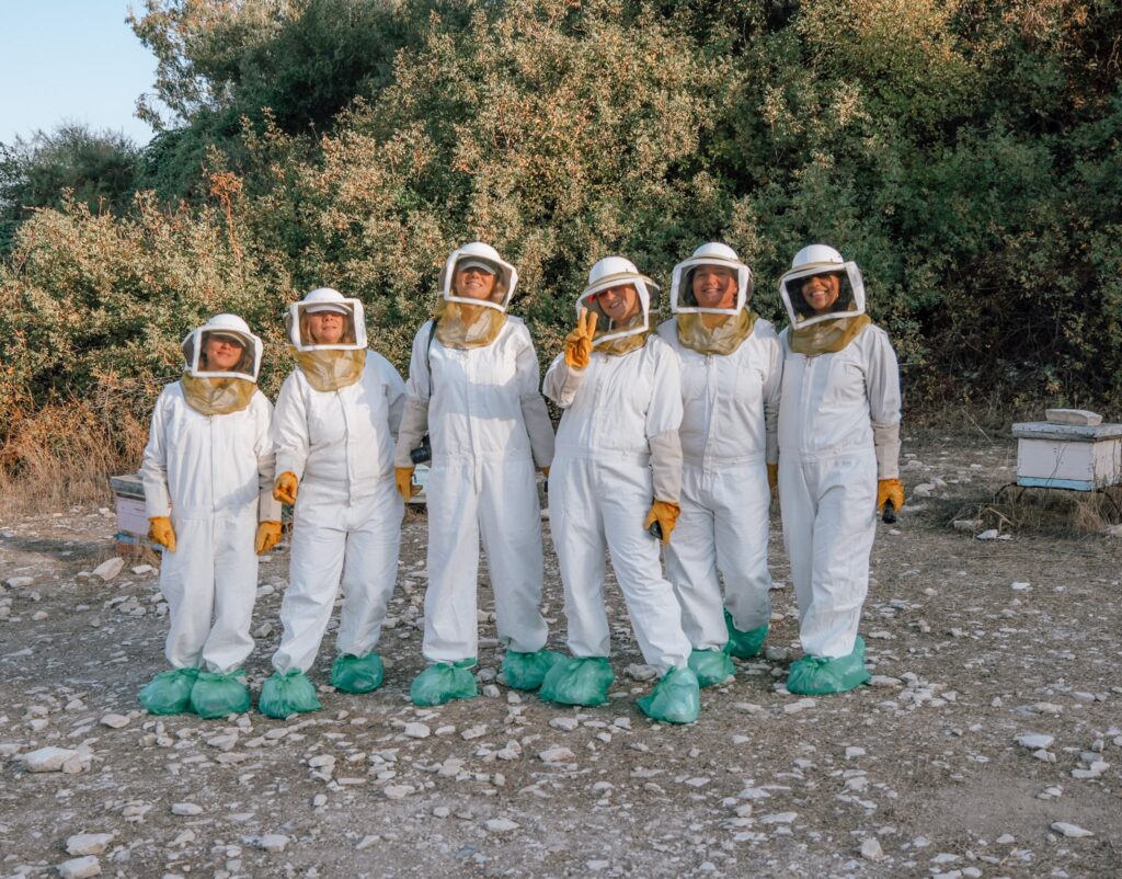 Six people wearing white beekeeping suits, gloves, and protective hats stand together on rocky ground surrounded by greenery and beehives. They pose closely, with some making peace signs.