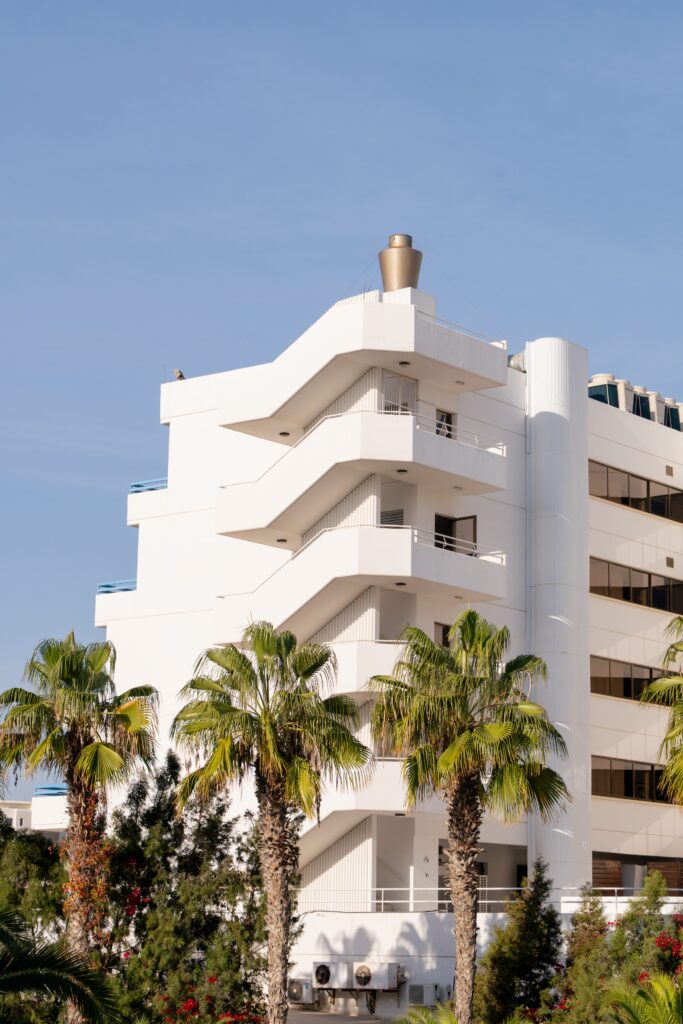 A white modern building with exterior staircases is seen behind tall palm trees on a sunny day, set against a clear blue sky.