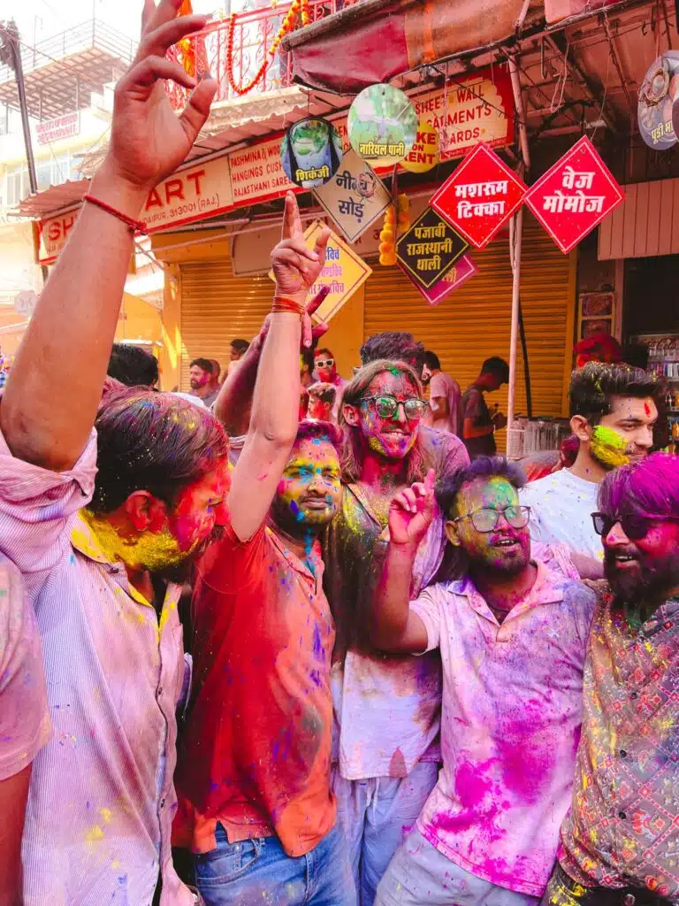A group of people covered in colorful powders celebrate Holi on a busy street, smiling and raising their hands in joy. Bright colors cover their faces and clothes, and shop signs in Hindi are visible in the background.