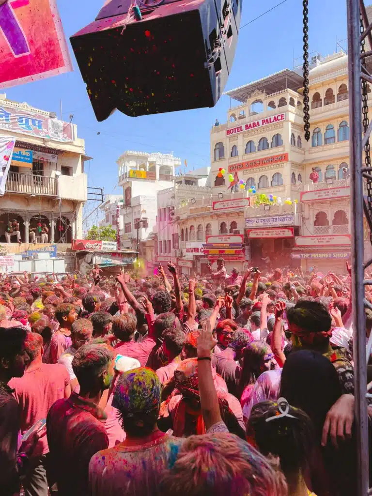 A large crowd celebrates Holi outdoors, covered in colorful powders. People raise their hands in joy. Historic buildings and banners are visible in the background under a clear blue sky.