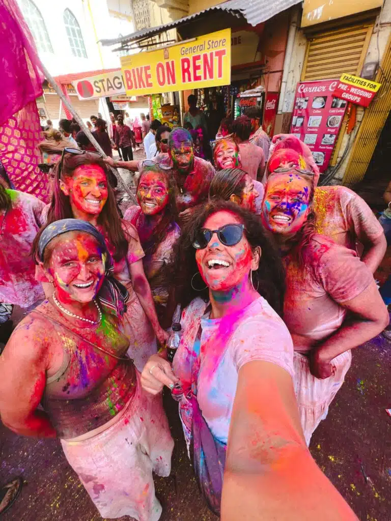 Backpacking India A group of people covered in colorful powder smile for a selfie during a street celebration, likely Holi festival. They appear joyful and are surrounded by vibrant colors with shops in the background.
