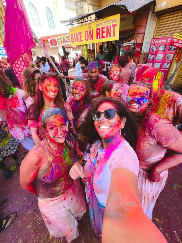 A group of people, smiling and covered in vibrant colored powders, celebrate Holi outdoors. They pose closely together for a selfie on a busy street with shops in the background.