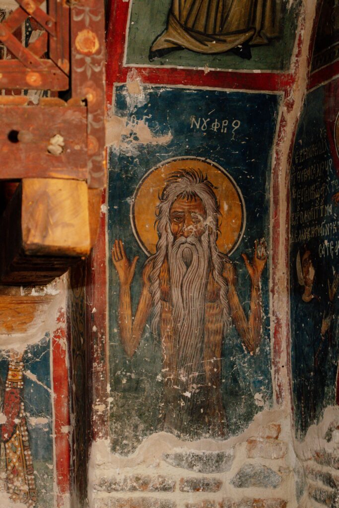 A religious fresco in Limassol, in winter, depicts an elderly bearded man with long hair, raised hands, and a golden halo against a blue background with Greek writing above. The aged artwork bears visible signs of time and wear.