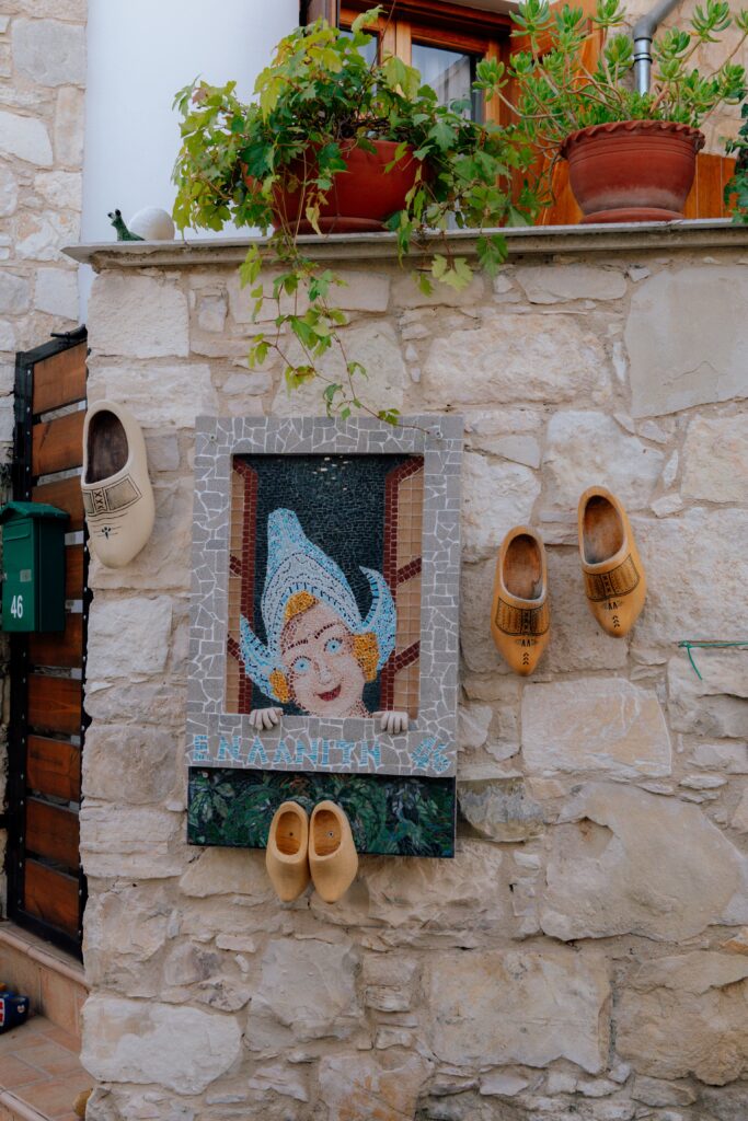 A mosaic portrait of a smiling person in a blue hat is mounted on a stone wall in Limassol in winter, surrounded by four wooden clogs and potted plants placed above the wall.