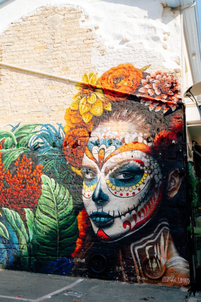 A vibrant mural features a woman's face painted in Day of the Dead style with colorful patterns and flowers in her hair, surrounded by lush green leaves against a textured brick wall.