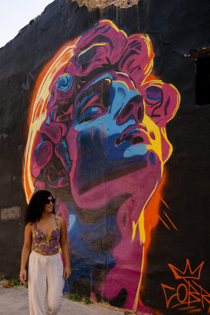 A woman in sunglasses stands next to a vibrant mural of a classical statue's head, painted in bold neon colors on a black wall, with Limassol in winter sunlight highlighting the artwork.