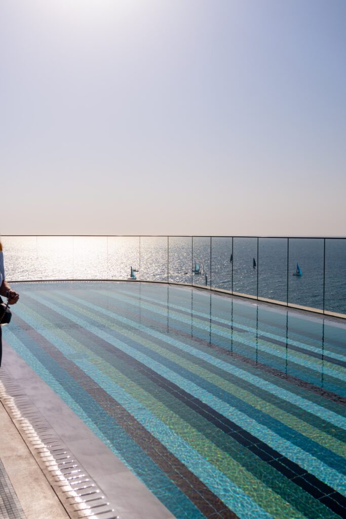 A rooftop swimming pool with striped tiles overlooks a calm, sunlit ocean. The sky is clear, glass barriers line the pool's edge, and part of a person is visible on the left side of the image.