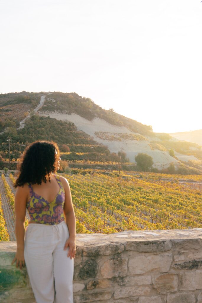 A woman with curly hair, wearing a floral top and light pants, leans against a stone wall overlooking a sunlit vineyard and rolling hills, capturing the tranquil charm of Limassol in winter.