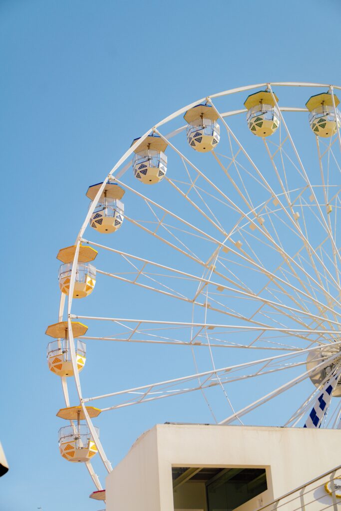 A large white Ferris wheel with yellow and white gondolas stands against a clear blue sky partially obscured by a white building at the bottom of the image.
