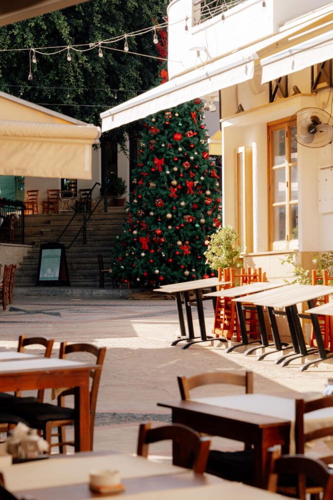 An outdoor café with empty tables and chairs basks in the sunlight. In the background, a large Christmas tree adorned with red ornaments and ribbons stands by a staircase next to a building—capturing the festive spirit of Limassol in winter.
