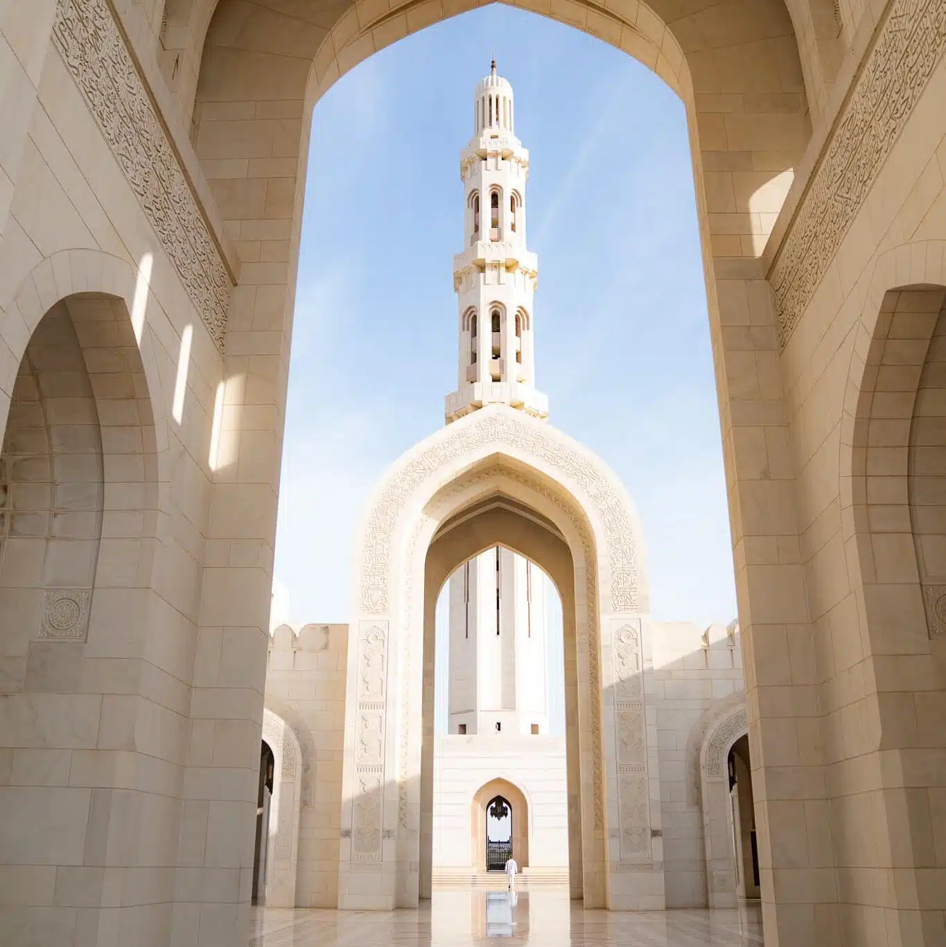 A tall minaret framed by ornate arches at the Sultan Qaboos Grand Mosque in Muscat, Oman, with sunlight illuminating the cream-colored stone—a reminder to consider What to Wear in Oman when visiting such revered sites.
