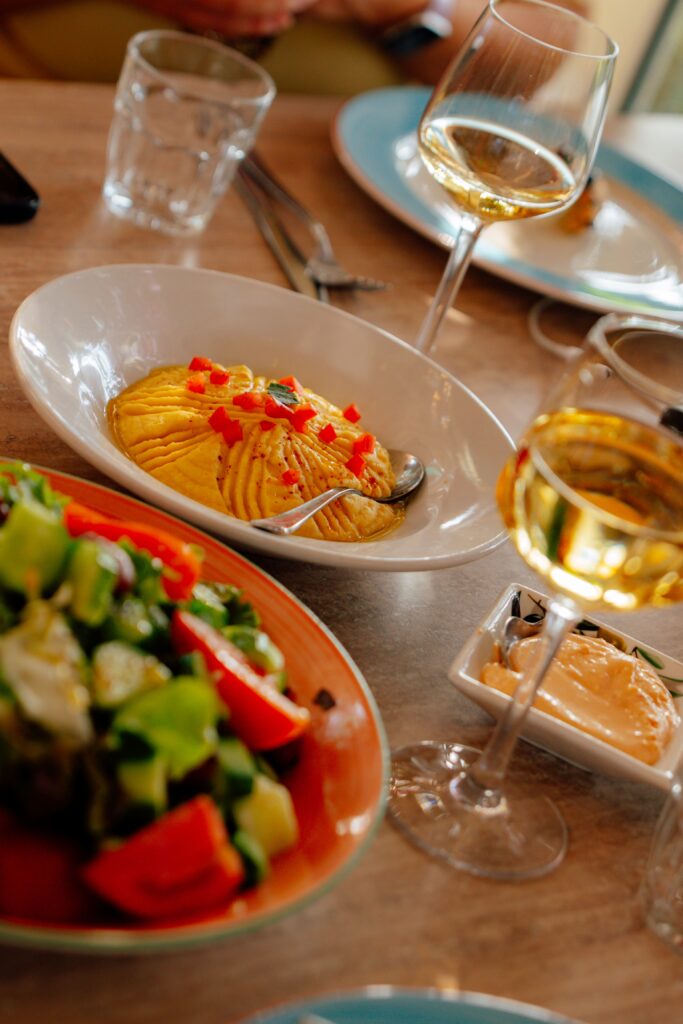 A table set with plates of hummus topped with diced red peppers, a fresh salad with cucumbers and tomatoes, glasses of white wine, and a glass of water—capturing the flavors of Limassol in winter. Cutlery and another plate are visible in the background.