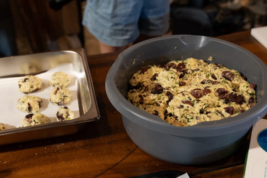 A large bowl of olive and herb bread dough sits on a wooden counter, next to a tray lined with parchment paper, holding several unbaked dough balls. A person in shorts stands in the background.