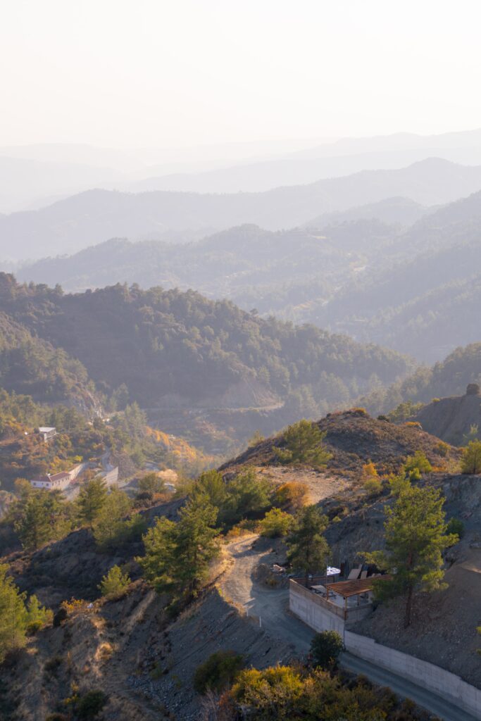 A scenic view of rolling mountains covered in trees, with a winding dirt road and a small building in the foreground, all beneath a hazy sky.