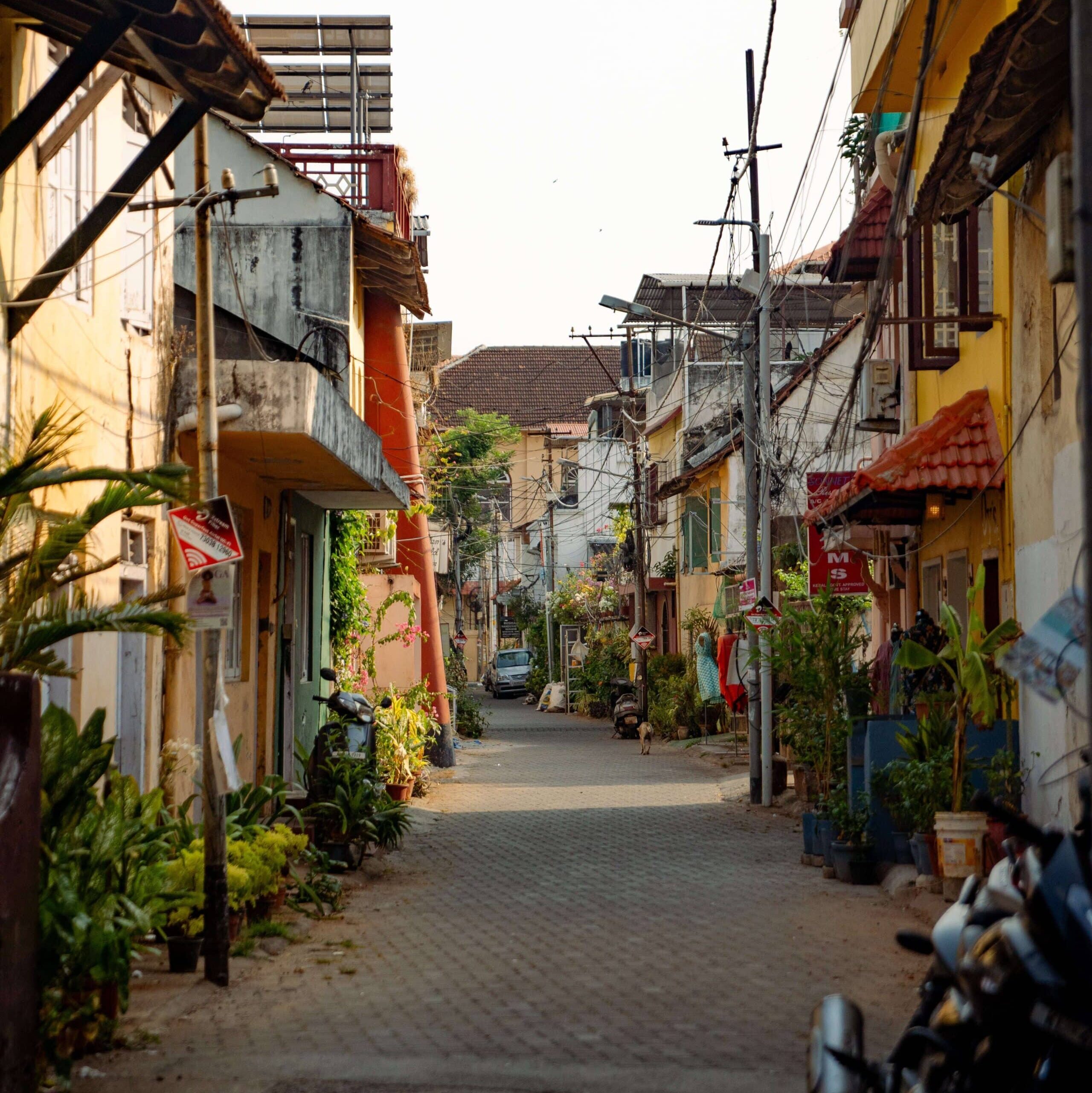 A narrow, sunlit street is lined with colorful, weathered buildings and outdoor plants. Power lines crisscross above, and a few parked motorcycles are visible. The scene feels peaceful and slightly rustic, with a blend of residential charm and urban decay.