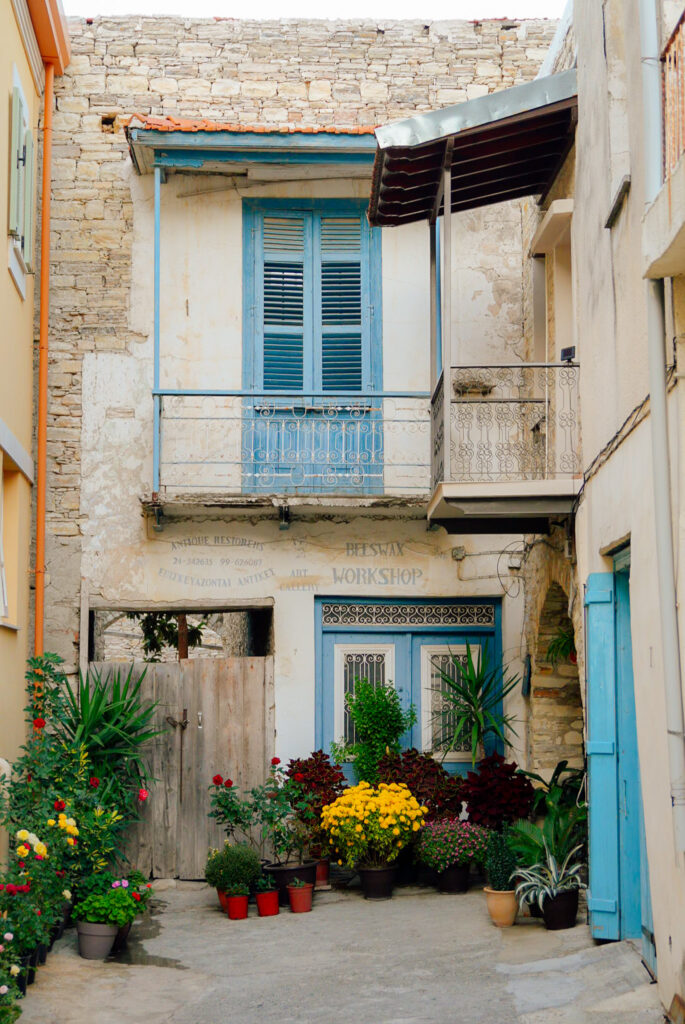 A rustic courtyard inspired by villages in Cyprus, with colorful potted plants and flowers, old stone walls, blue wooden doors and shutters, and a balcony with ornate railing above the entrance.