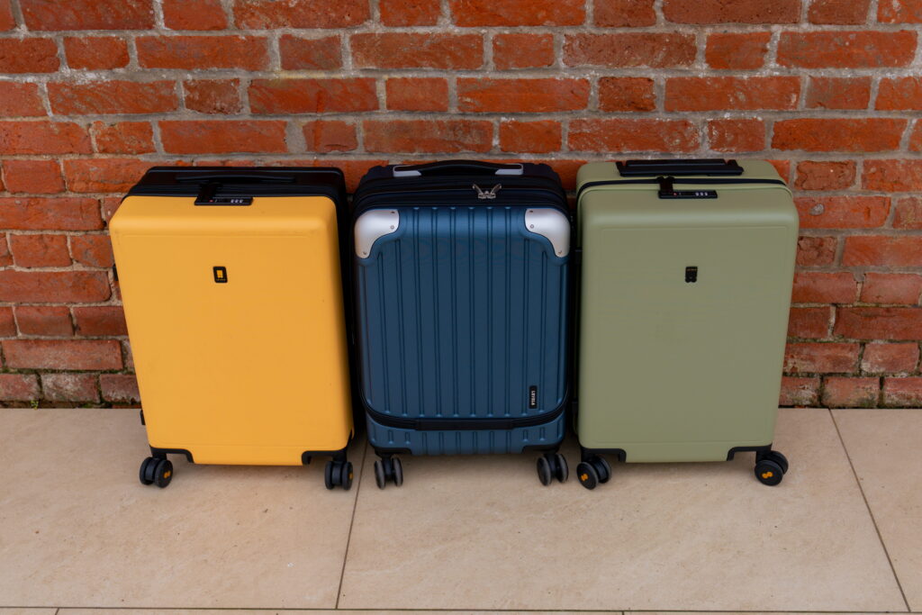 Three hard-shell suitcases—yellow, blue, and green—stand upright side by side on a tiled floor in front of a red brick wall. Each suitcase, including the sleek Level8 carry-on, features four wheels and a retractable handle.