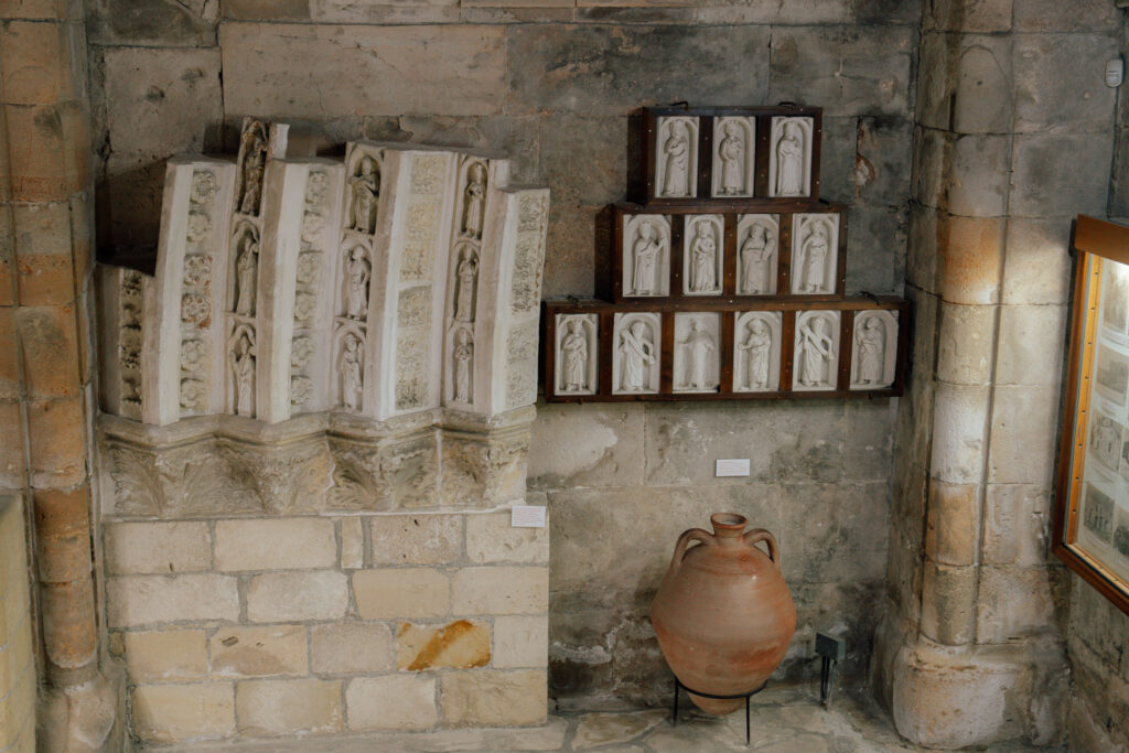 A stone wall with carved stone reliefs of figures on two shelves, a large clay amphora on the floor, and a glass display case on the right—an intriguing museum exhibit perfect to explore during one day in Limassol.