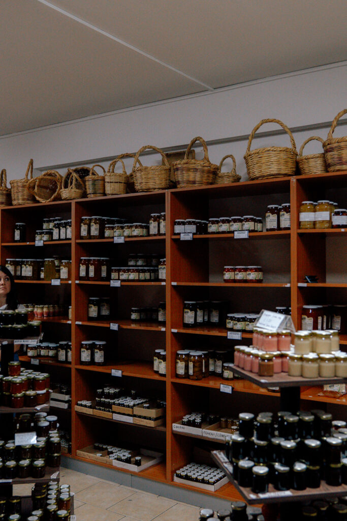Wooden shelves filled with jars of preserves and jams are arranged against a wall, with woven baskets displayed on top—reminiscent of markets found in villages in Cyprus. A rotating stand holds more jars in the foreground inside this specialty food store.
