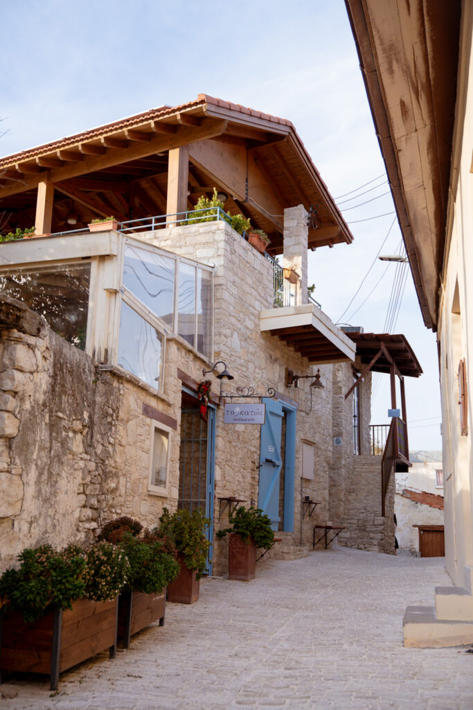 A narrow stone alleyway typical of villages in Cyprus, lined with potted plants and rustic buildings, including a two-story stone house with blue doors, balconies, and a wooden roof, under a clear sky.
