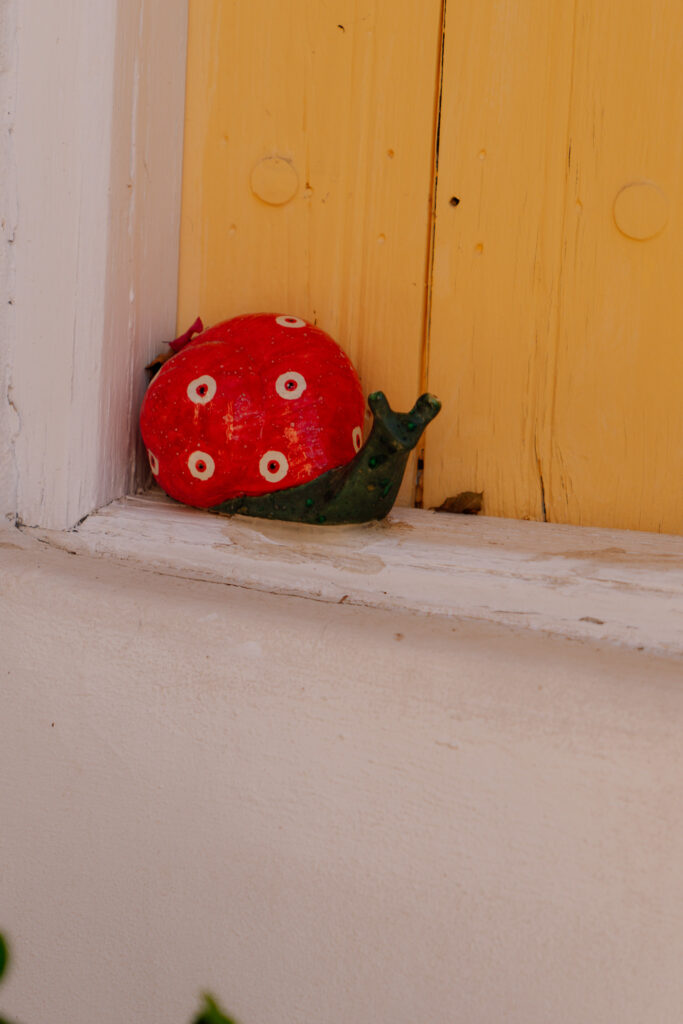 A small decorative snail sculpture with a red shell and white spots sits on a white windowsill beneath a yellow shutter, bringing a touch of the charm found in villages in Cyprus.