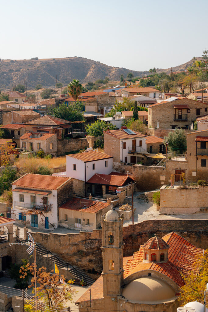A hillside Mediterranean village with stone houses, red-tiled roofs, and a church tower, reminiscent of villages in Cyprus. The dry landscape features scattered trees and rolling hills under a clear sky.