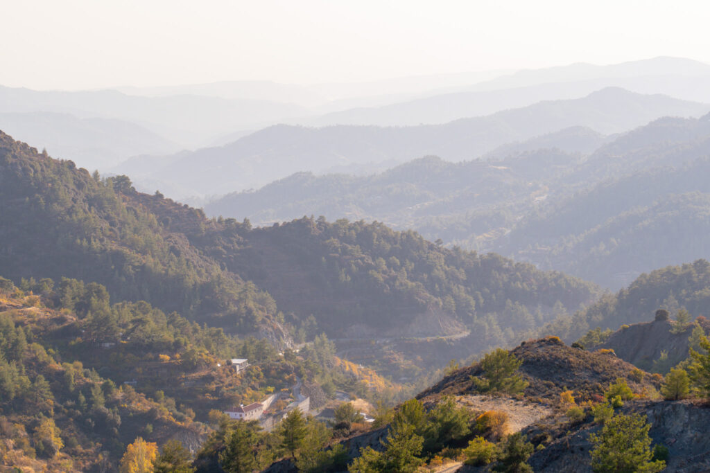 Mountain landscape with multiple tree-covered ridges fading into the misty distance, bathed in soft golden sunlight. Small houses, reminiscent of mountain villages in Cyprus, are nestled among the trees in the lower part of the scene.