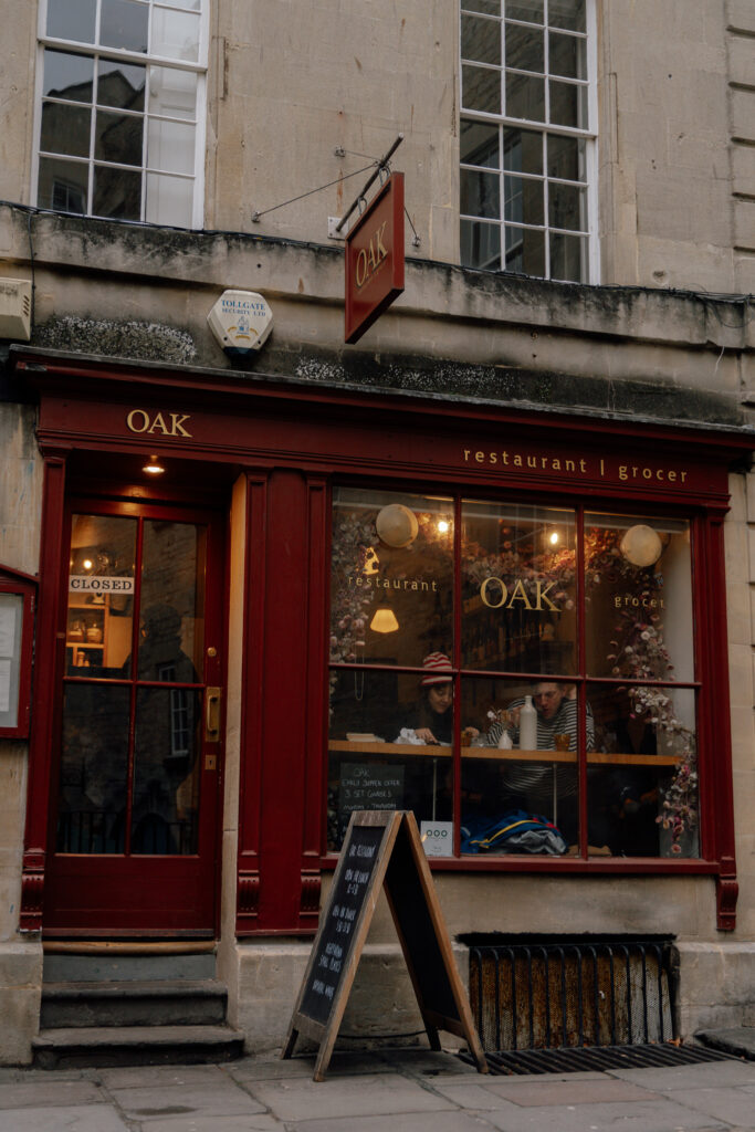 A cozy restaurant and grocer called "OAK" with a red storefront, warm lights inside, and people sitting by the window—one of the charming photo spots in Bath. A chalkboard menu sits on the sidewalk outside the entrance.