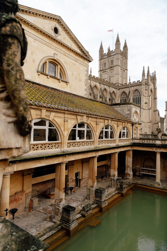 A must-see during one day in Bath: the historic Roman Baths with arched windows, columns, and greenish water, framed by the towering Gothic spires of Bath Abbey under a cloudy sky.