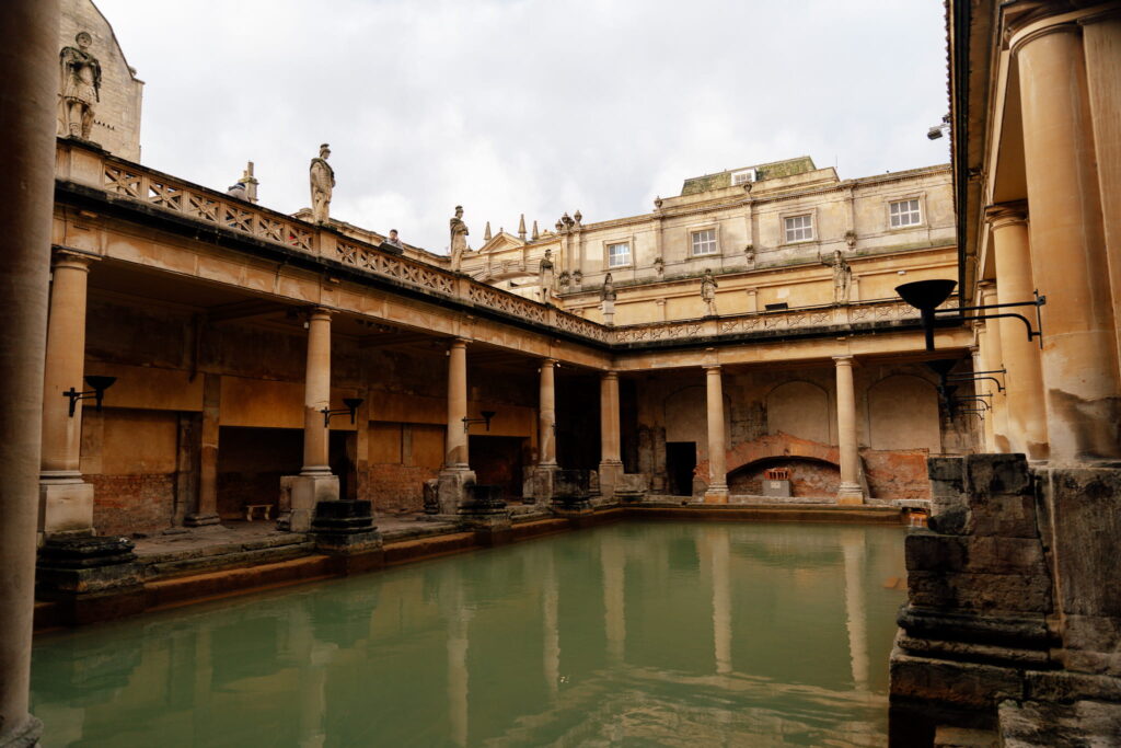Experience the ancient Roman bath in Bath, where tall columns and statues rise above still greenish water—an iconic sight to explore during one day in Bath under a moody, cloud-filled sky.