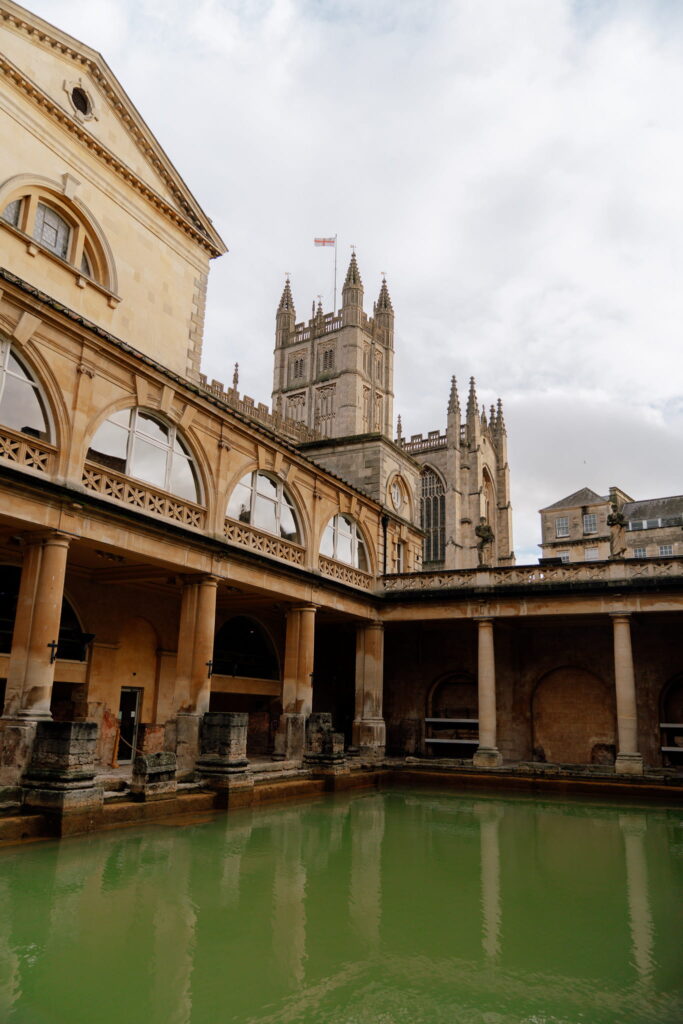 The image shows the Roman Baths in Bath, England, with arched columns around a green pool—an iconic stop for one day in Bath. In the background, Bath Abbey’s tall, intricate Gothic towers rise against a cloudy sky.