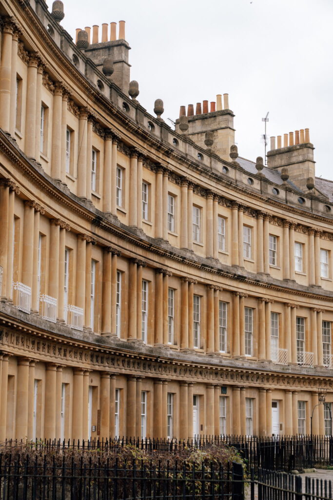 A curved row of Georgian-style terraced houses with tall chimneys, cream-colored stone facades, and black iron railings—an iconic sight for anyone enjoying one day in Bath under an overcast sky.