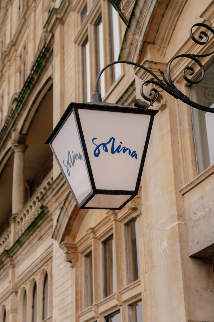 A white lantern-style street sign with the word "Solina" written in blue cursive hangs on an ornate metal bracket, evoking the charm you’d discover during one day in Bath, against a beige stone building with arched windows.