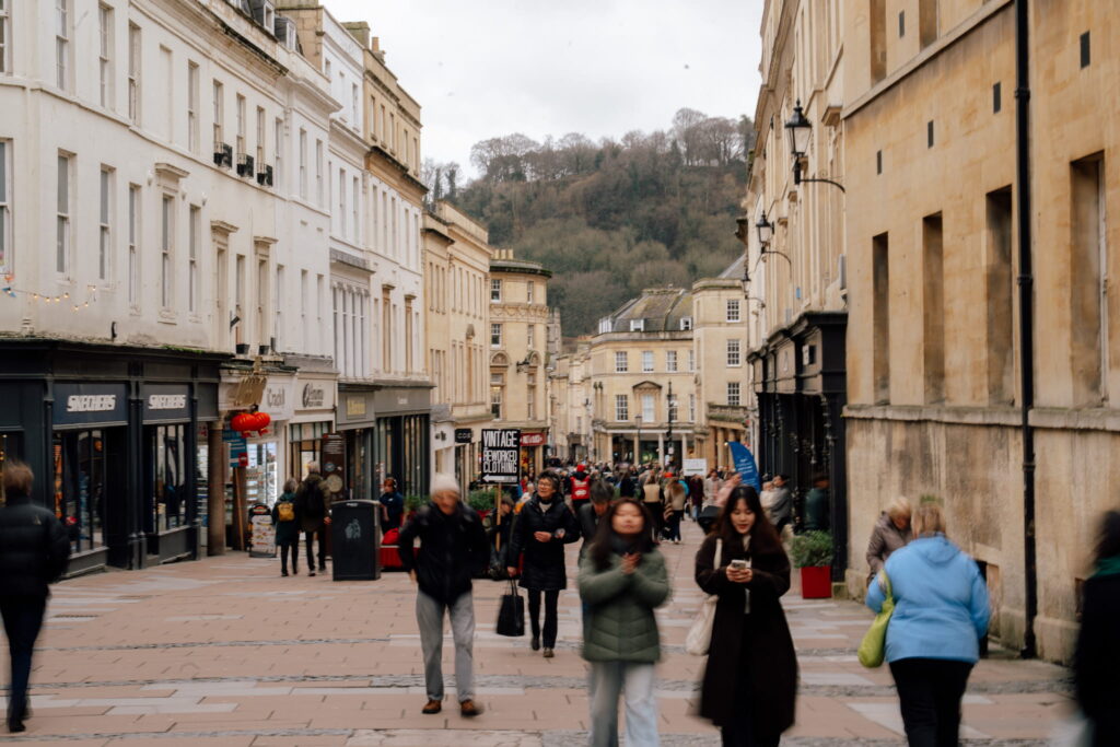 A busy pedestrian street in Bath with people walking and shopping, lined with historic beige buildings. Trees and hills are visible in the background under a cloudy sky—a perfect scene to experience during one day in Bath.