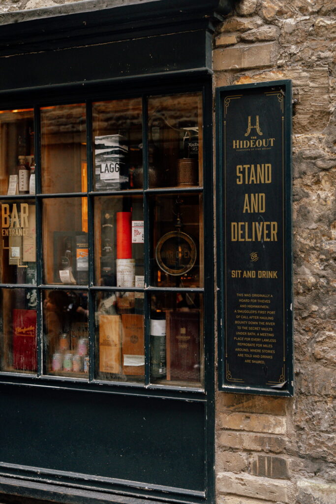 A rustic bar exterior with a window displaying bottles and a black sign reading “Hideout Stand and Deliver, Sit and Drink” mounted on a stone wall—a cozy, vintage spot perfect to discover during one day in Bath.