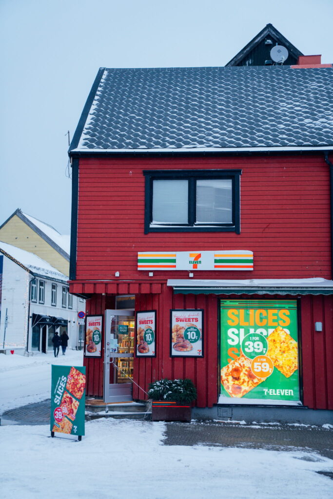 A red 7-Eleven store in a snowy area, with posters advertising pizza slices and sweets on the windows—an ideal snack stop on your 3 Day Tromso Itinerary. Other buildings and a few people are visible in the background.