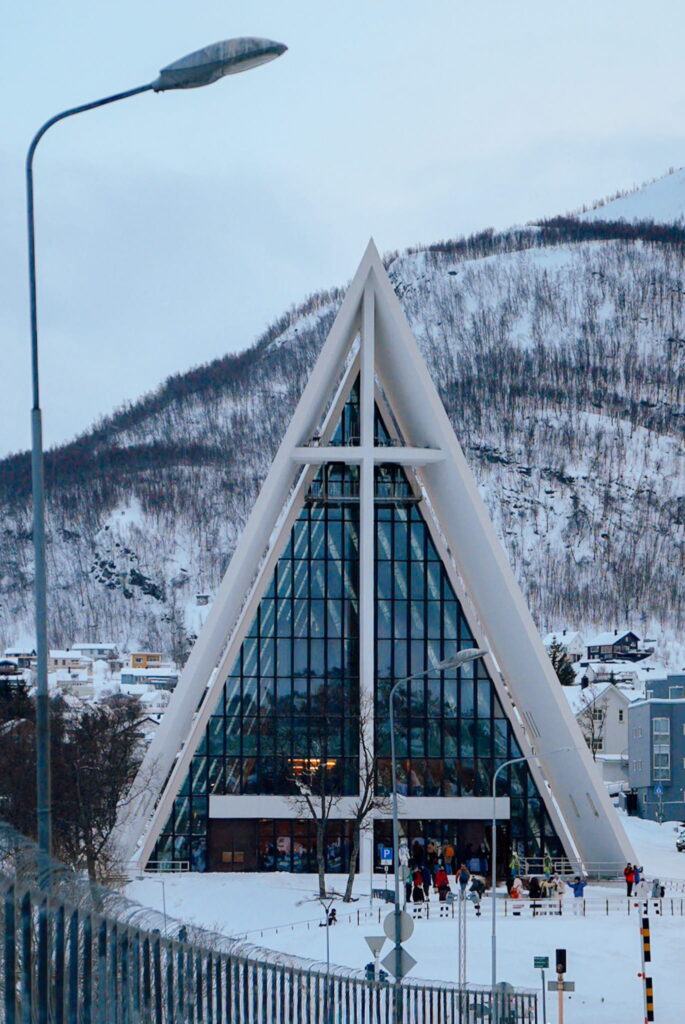 A modern, triangular glass and white concrete building stands in a snowy landscape with bare trees and hills—an architectural highlight you’ll spot on a 3 Day Tromso Itinerary. Several people gather at its entrance beneath streetlights.