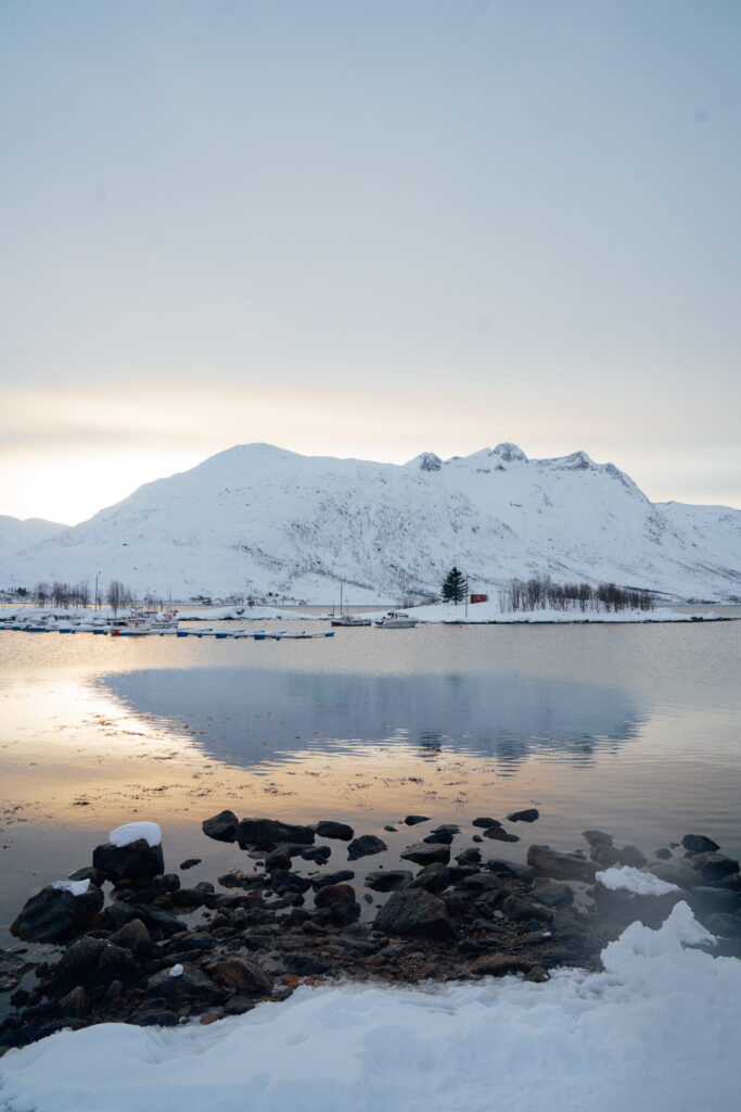 Snowy mountains reflect in a calm lake at sunset, with a rocky shoreline in the foreground—an ideal scene to include in your 3 Day Tromso Itinerary as soft, warm light illuminates the sky and water.