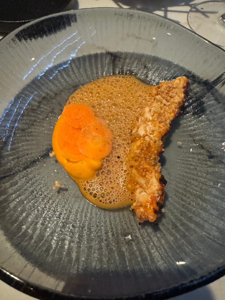 A gray plate with a breaded, fried fish fillet, bright orange sauce, and thinly sliced carrots, garnished with light brown foam—perfect for savoring after a day of adventure on your 3 Day Tromso Itinerary.