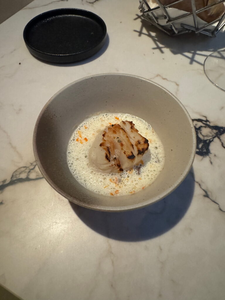 A bowl with a piece of grilled seafood, possibly a scallop, sits in a light, foamy sauce on a marble table—an elegant dish you might savor during your 3 Day Tromso Itinerary. A dark plate, basket, and part of a wine glass appear in the background.
