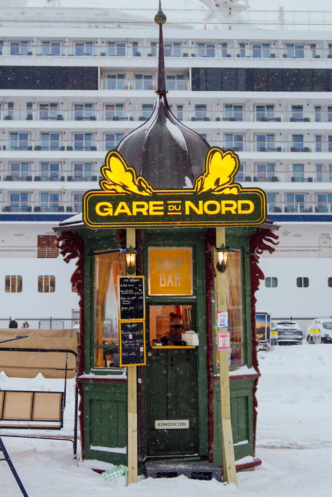A small green kiosk labeled "Gare du Nord" with a yellow neon sign sits in a snowy setting, perfect for your 3 Day Tromso Itinerary. Inside, a person serves at a soup bar while a cruise ship glides by in the background.