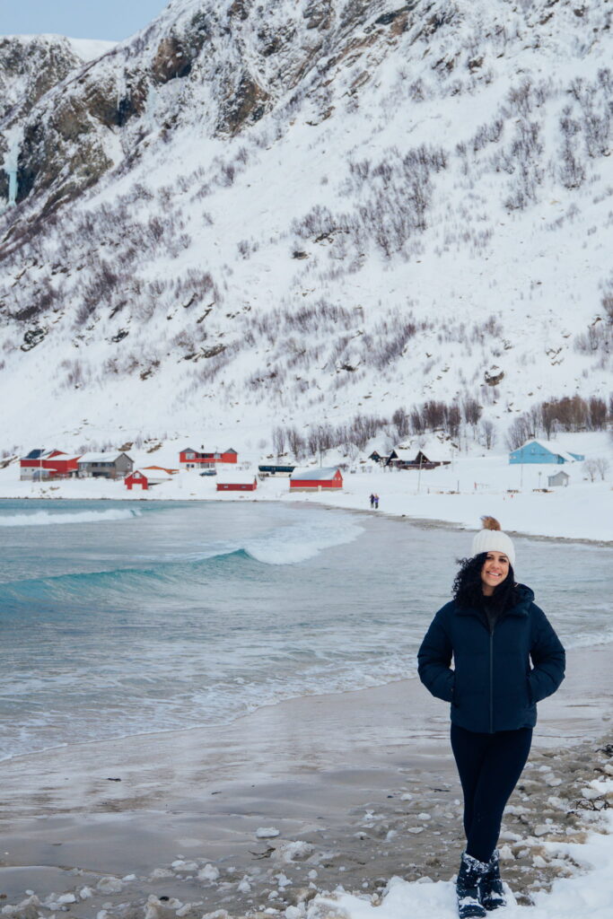 A woman in a dark winter coat stands on a snowy beach beside the sea, framed by snow-covered mountains and colorful houses—a scene perfect for your 3 Day Tromso Itinerary.