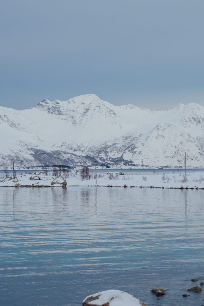 Snow-covered mountains with rugged peaks reflect in a calm body of water under a cloudy sky—an idyllic scene perfect for your 3 Day Tromso Itinerary. The snowy shoreline and bare trees create a serene winter landscape.
