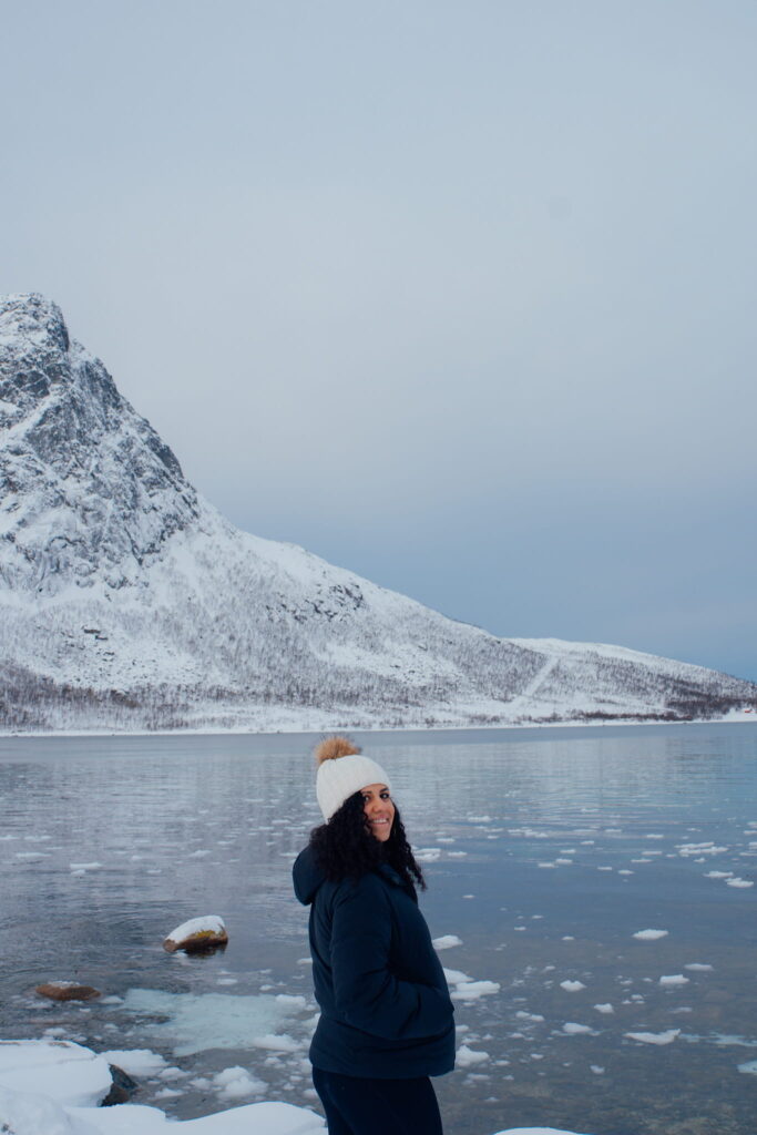A person in a dark winter coat and white beanie stands by a snowy lakeshore, smiling, as if capturing the perfect moment for a 3 Day Tromso Itinerary, with snow-covered mountains and a calm, partially frozen lake in the background.